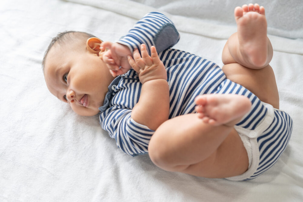 A baby lies on a soft surface, lifting legs and bringing hands toward the face.