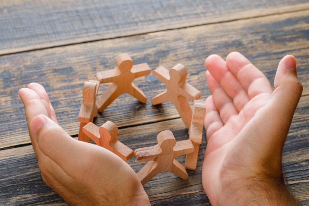 Hands cupping a circle of small wooden human figures on a wooden surface.