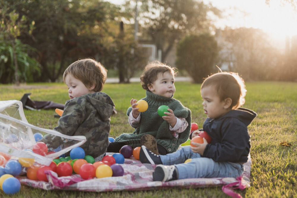 Three young children sit on a blanket outdoors, playing with colourful plastic balls in a grassy garden.