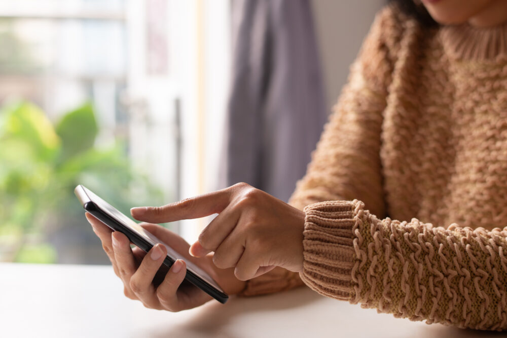 A person in a textured sweater uses a smartphone while sitting near a window.