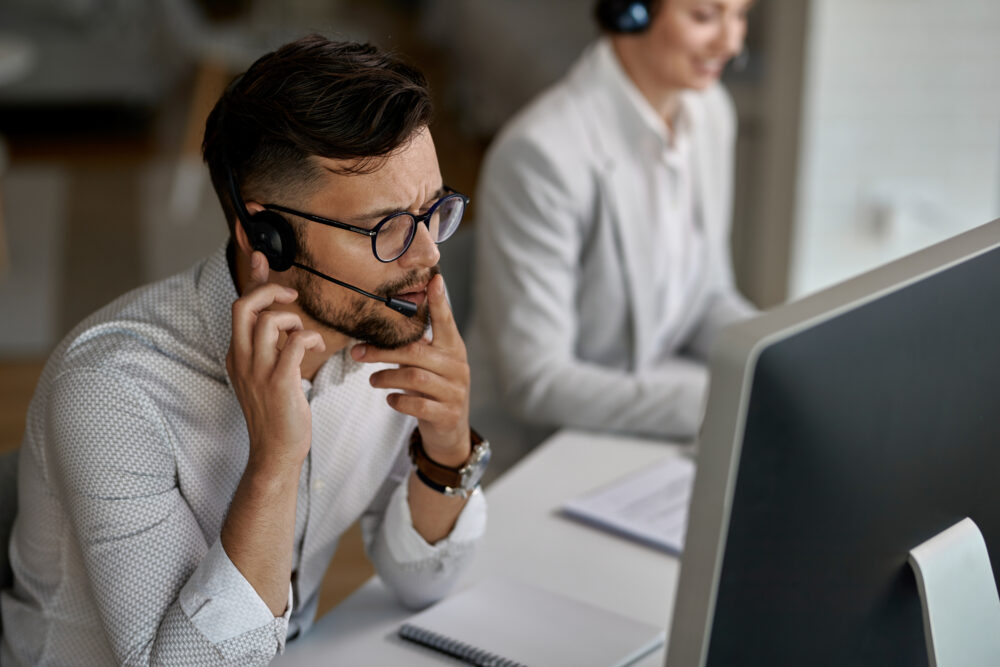 A person wearing a headset sits at a desk using a computer while another person works in the background.