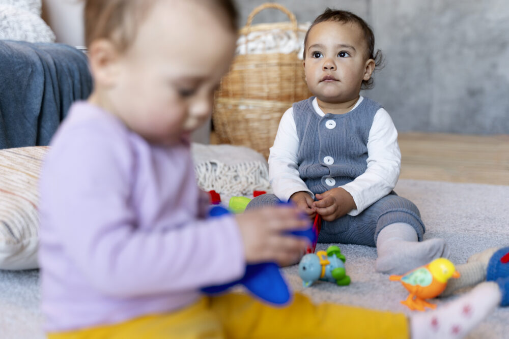 Two young children sit on a carpet playing with colourful toys, with one in the foreground and one sitting upright in the background.