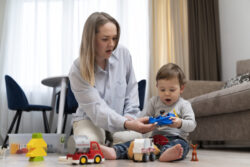 An adult sits on the floor helping a child play with toy animals and vehicles in a living room.