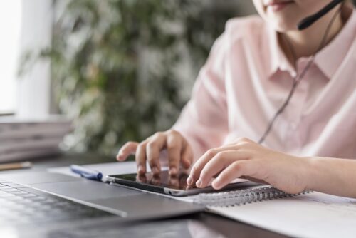 Hands using a tablet on a desk beside a laptop and notebook, with a headset visible.
