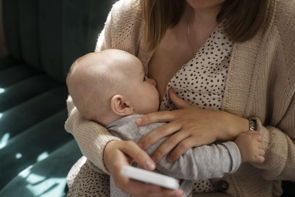 A baby is held and breastfed by an adult, with one hand holding a phone.
