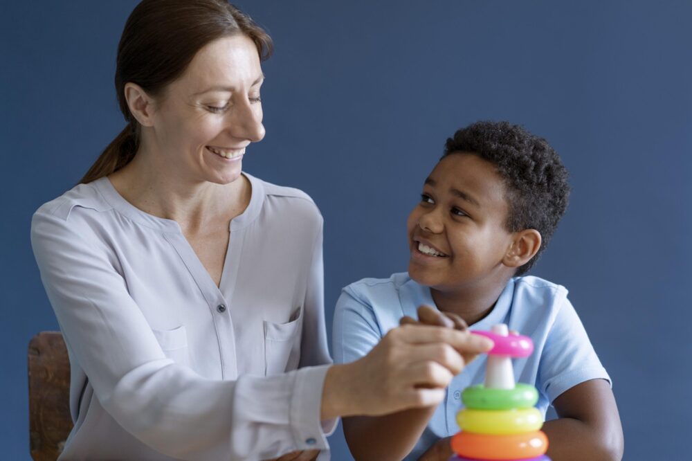 An adult and a child sit together at a table placing colourful stacking rings on a toy.