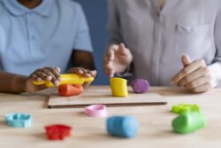 Hands use a rolling pin to shape colourful modelling clay on a table alongside cutters.