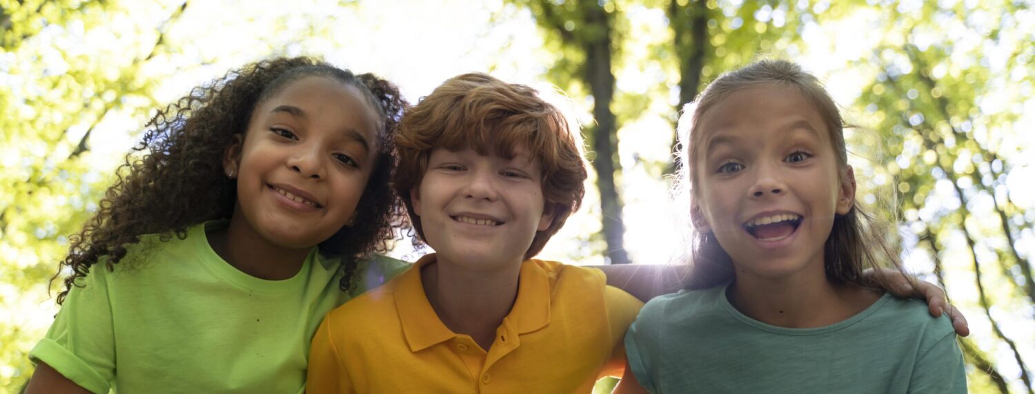 Three children stand close together outdoors with trees and sunlight in the background.