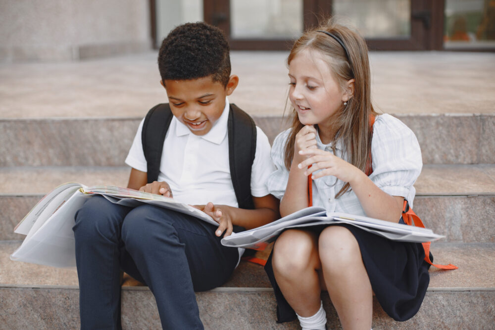 Two schoolchildren sit on steps reading books together with backpacks beside them.