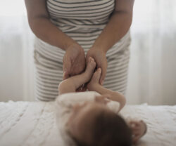 Hands gently hold and move a baby’s legs while the baby lies on a soft surface.