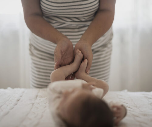 Hands gently hold and move a baby’s legs while the baby lies on a soft surface.