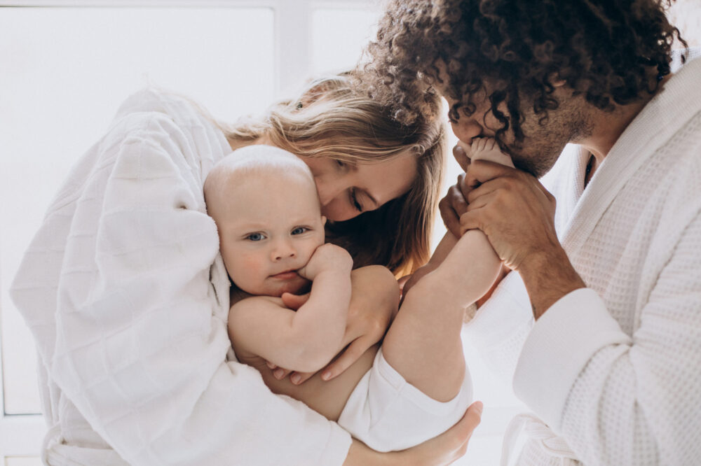 Two adults hold a baby close, with one gently holding the baby’s foot near their face.