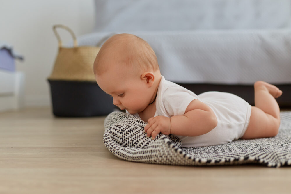 A baby lies on a mat on the floor, lifting the head while resting on the arms.