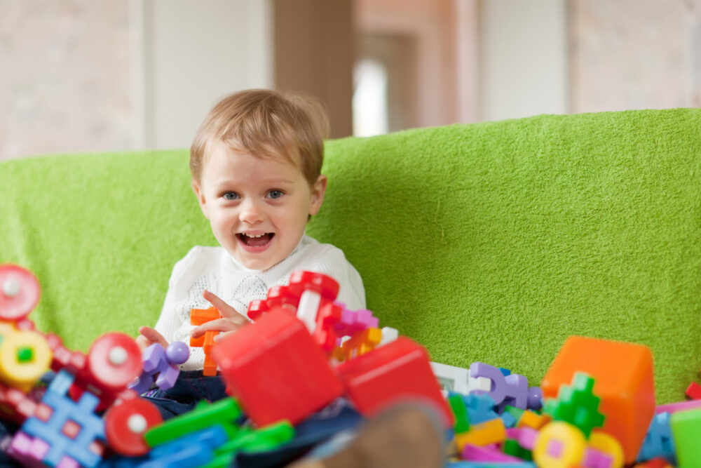 A young child sits on a sofa surrounded by colourful toy blocks and play items.