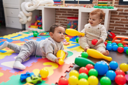 Two babies sit on a foam play mat surrounded by colourful balls and toys, holding yellow bowling‑pin toys.