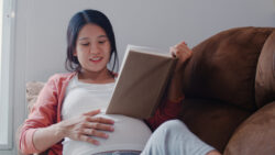 A pregnant person sits on a sofa reading a book while holding their belly.