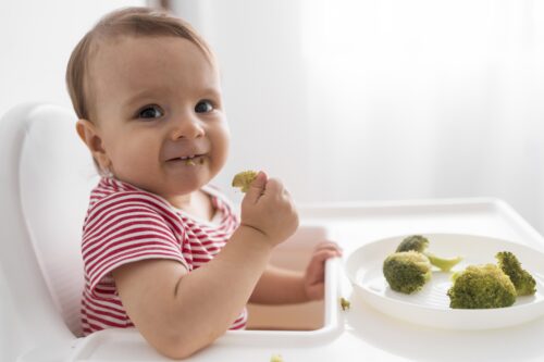 A baby sits in a highchair eating broccoli from a plate.