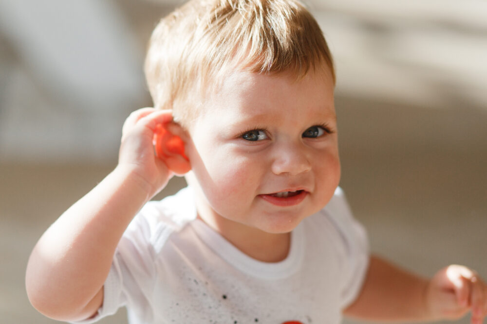 A baby holds a small red object to one ear, as if listening.