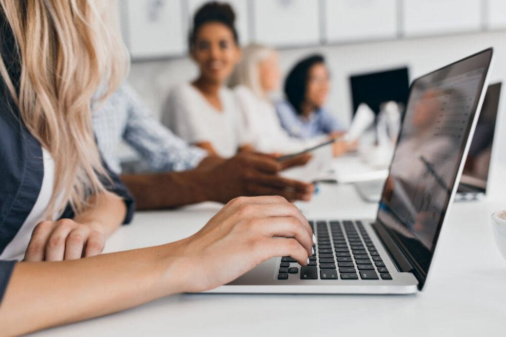 Hands type on a laptop at a desk while other people sit and discuss in the background.