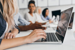 Hands type on a laptop at a desk while other people sit and discuss in the background.