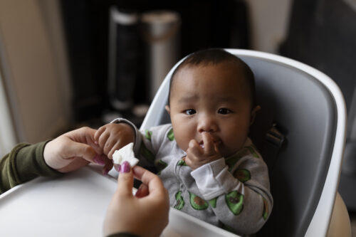 A baby sits in a high chair while an adult holds a spoon and feeds them.
