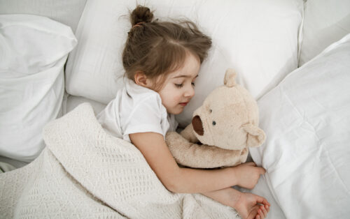 A child lies in bed hugging a teddy bear under a blanket.