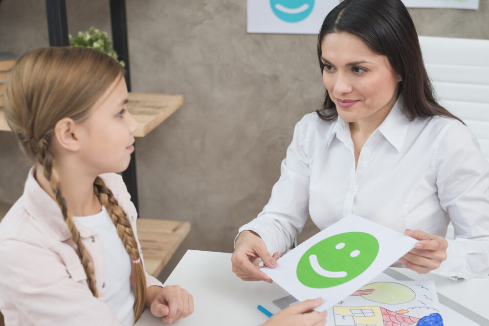 An adult shows a green smiley‑face card to a child during an activity at a table.