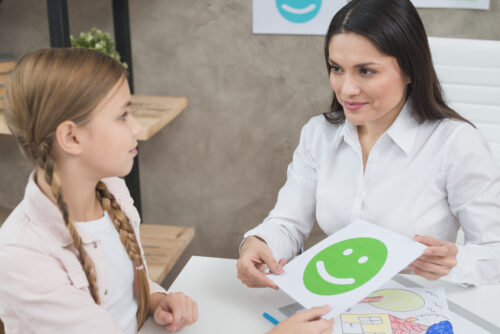 An adult shows a green smiley‑face card to a child during an activity at a table.