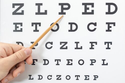 Close-up of an eye chart with black letters and a hand pointing at the top row with a wooden pointer.