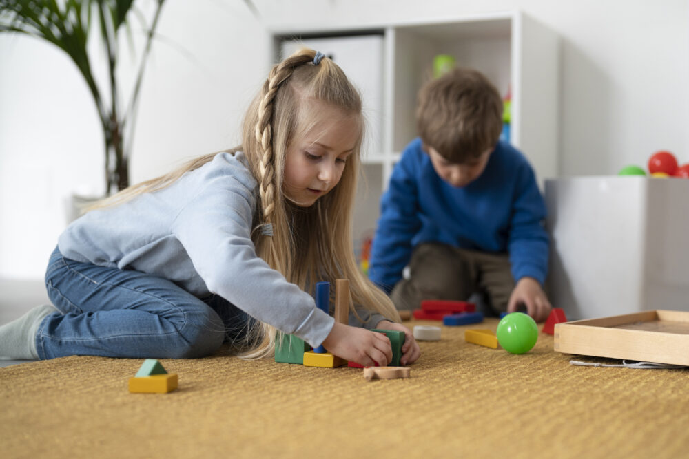 Two children sit on the floor playing with colourful wooden blocks and shapes.