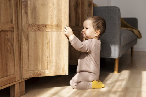 A young child kneels on the floor, opening a wooden cabinet door indoors.