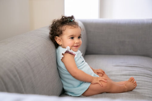A baby sits upright on a grey sofa with legs stretched out, wearing a light blue outfit.