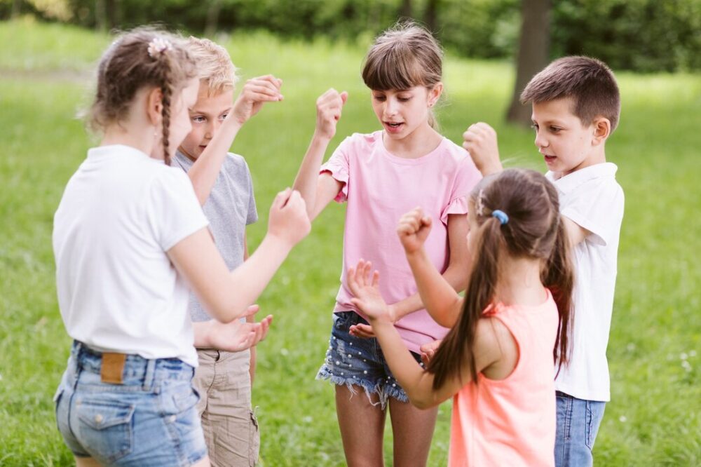 Five children stand in a circle outdoors, playing a hand game together on the grass