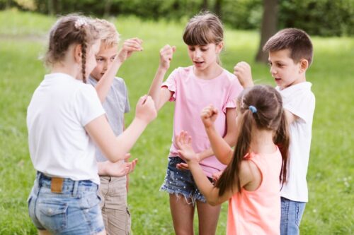 Five children stand in a circle outdoors, playing a hand game together on the grass