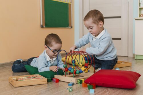 Two young children sit on the floor, playing with a bead maze toy and blocks.
