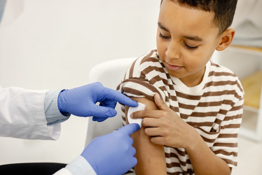 A healthcare worker wearing gloves places a cotton pad on a child’s upper arm after an injection.