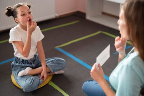 An adult holds up a card while a child sits on the floor practising mouth movements during a speech activity.