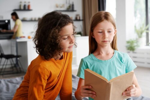 Two children sit indoors, with one reading a book while the other looks on.
