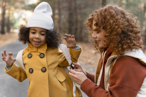 An adult fastens a child’s backpack strap while standing together on a tree‑lined path.