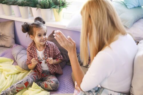 A child sits on a bed facing an adult, raising hands for a clapping game indoors.